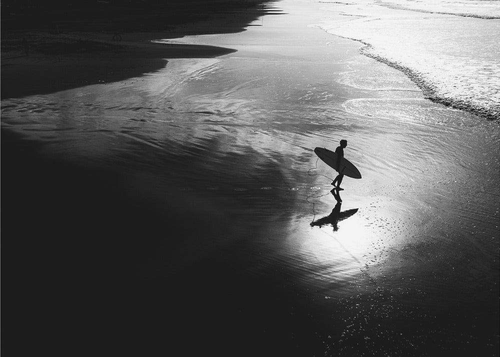 A high-contrast, black and white photograph of a silhouetted surfer carrying a surfboard and walking along the wet, reflective sand at the water's edge. Decor