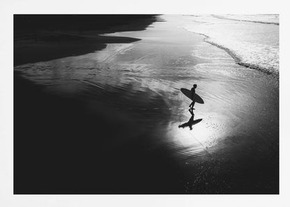 A high-contrast, black and white photograph of a silhouetted surfer carrying a surfboard and walking along the wet, reflective sand at the water's edge. Decor