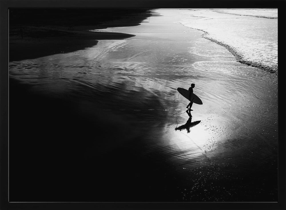 A high-contrast, black and white photograph of a silhouetted surfer carrying a surfboard and walking along the wet, reflective sand at the water's edge. Decor