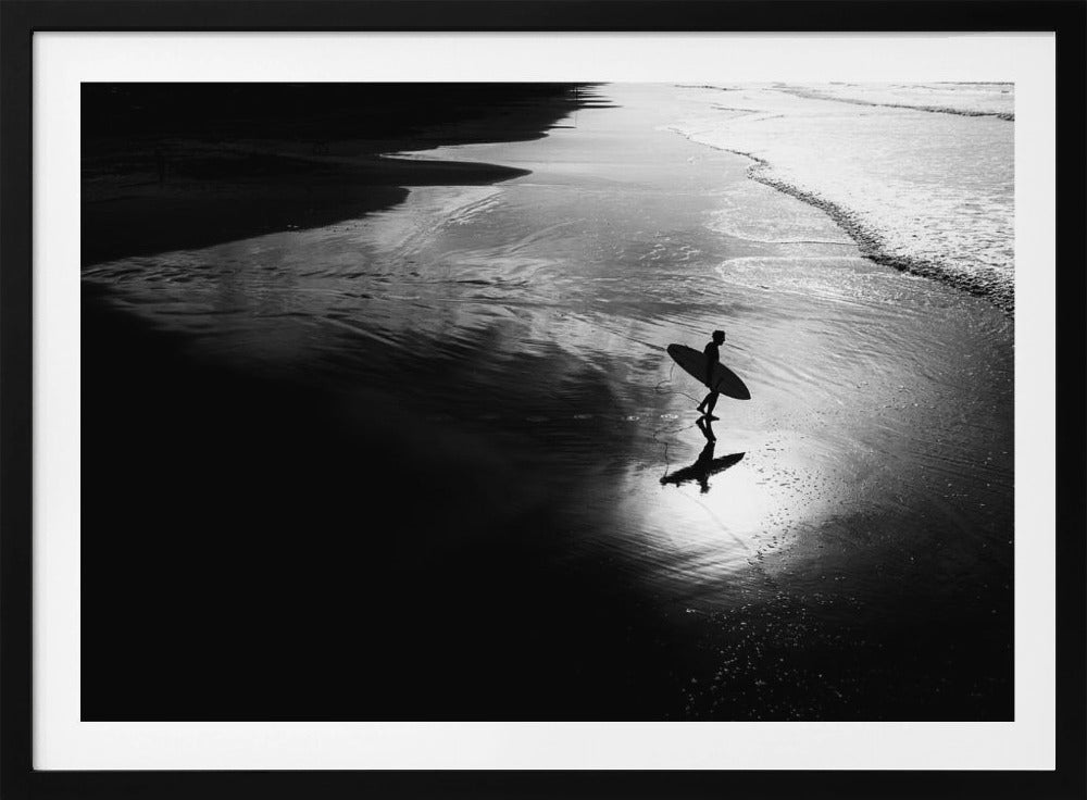 A high-contrast, black and white photograph of a silhouetted surfer carrying a surfboard and walking along the wet, reflective sand at the water's edge. Decor