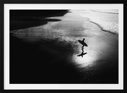 A high-contrast, black and white photograph of a silhouetted surfer carrying a surfboard and walking along the wet, reflective sand at the water's edge. Decor