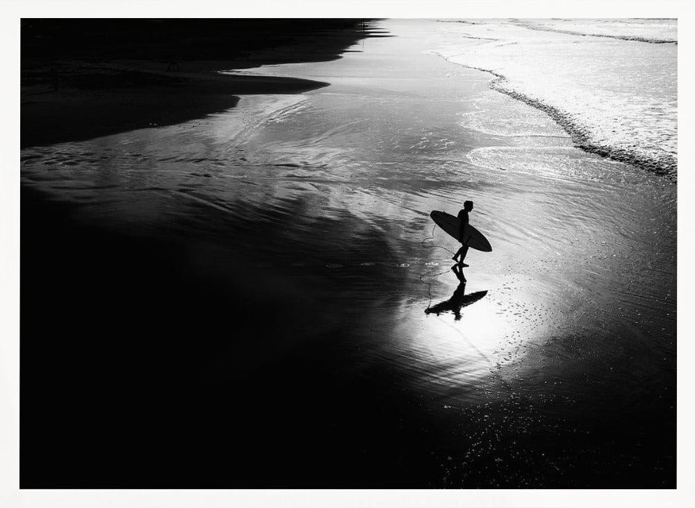 A high-contrast, black and white photograph of a silhouetted surfer carrying a surfboard and walking along the wet, reflective sand at the water's edge. Decor