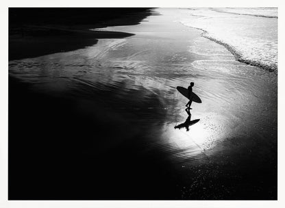A high-contrast, black and white photograph of a silhouetted surfer carrying a surfboard and walking along the wet, reflective sand at the water's edge. Decor