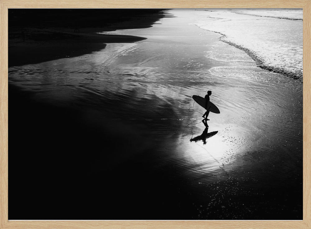 A high-contrast, black and white photograph of a silhouetted surfer carrying a surfboard and walking along the wet, reflective sand at the water's edge. Decor