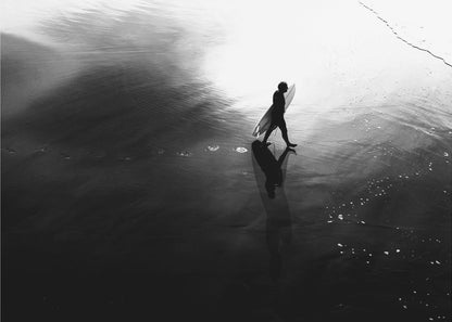A high-contrast, black and white photograph of a surfer silhouetted against the bright reflection of the sun on the water. The surfer is carrying a surfboard and walking along the wet sand, leaving footprints behind. Their long shadow is cast in front of them on the dark, glistening sand. Print