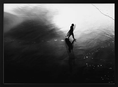 A high-contrast, black and white photograph of a surfer silhouetted against the bright reflection of the sun on the water. The surfer is carrying a surfboard and walking along the wet sand, leaving footprints behind. Their long shadow is cast in front of them on the dark, glistening sand. Print