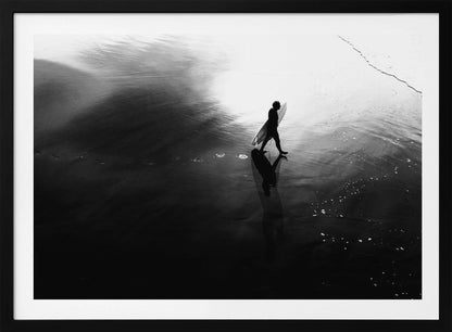 A high-contrast, black and white photograph of a surfer silhouetted against the bright reflection of the sun on the water. The surfer is carrying a surfboard and walking along the wet sand, leaving footprints behind. Their long shadow is cast in front of them on the dark, glistening sand. Print