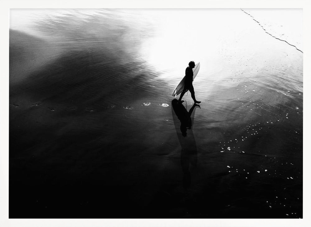 A high-contrast, black and white photograph of a surfer silhouetted against the bright reflection of the sun on the water. The surfer is carrying a surfboard and walking along the wet sand, leaving footprints behind. Their long shadow is cast in front of them on the dark, glistening sand. Print