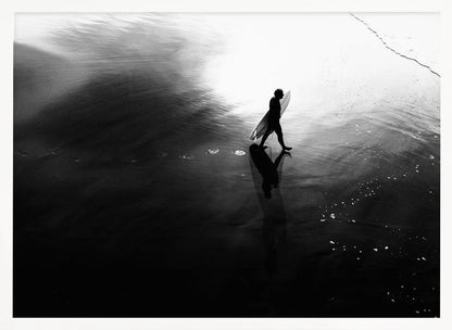 A high-contrast, black and white photograph of a surfer silhouetted against the bright reflection of the sun on the water. The surfer is carrying a surfboard and walking along the wet sand, leaving footprints behind. Their long shadow is cast in front of them on the dark, glistening sand. Print