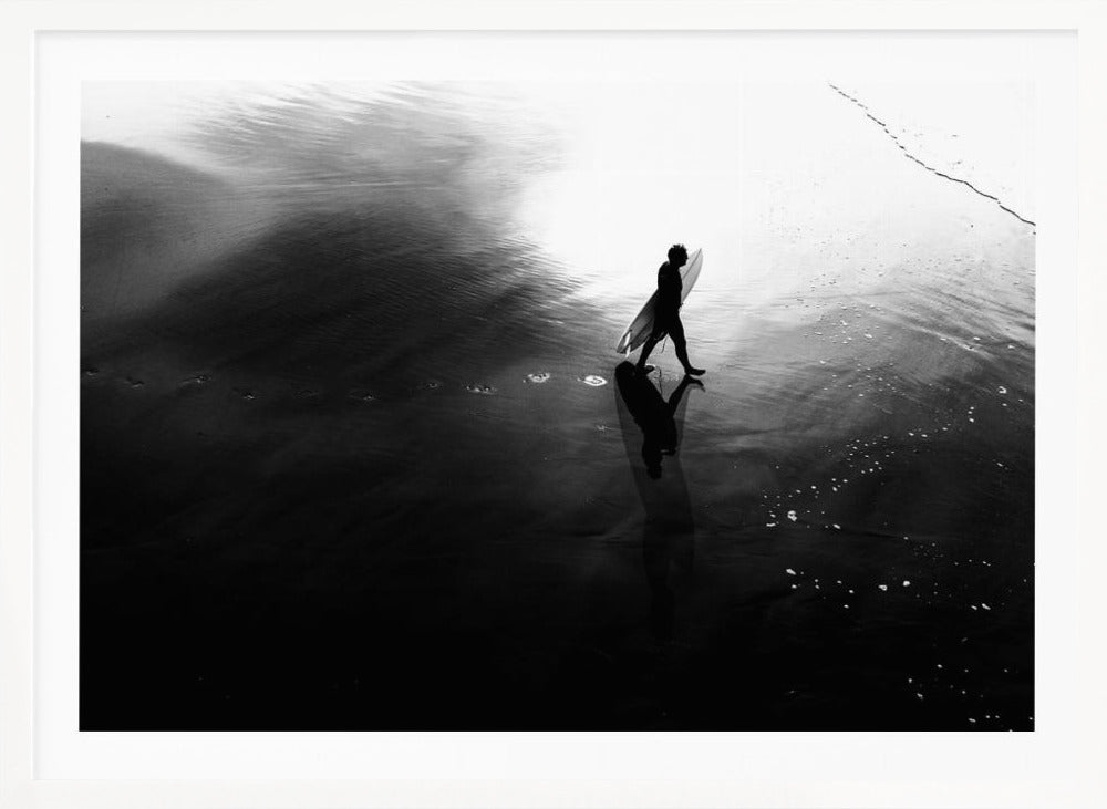 A high-contrast, black and white photograph of a surfer silhouetted against the bright reflection of the sun on the water. The surfer is carrying a surfboard and walking along the wet sand, leaving footprints behind. Their long shadow is cast in front of them on the dark, glistening sand. Print