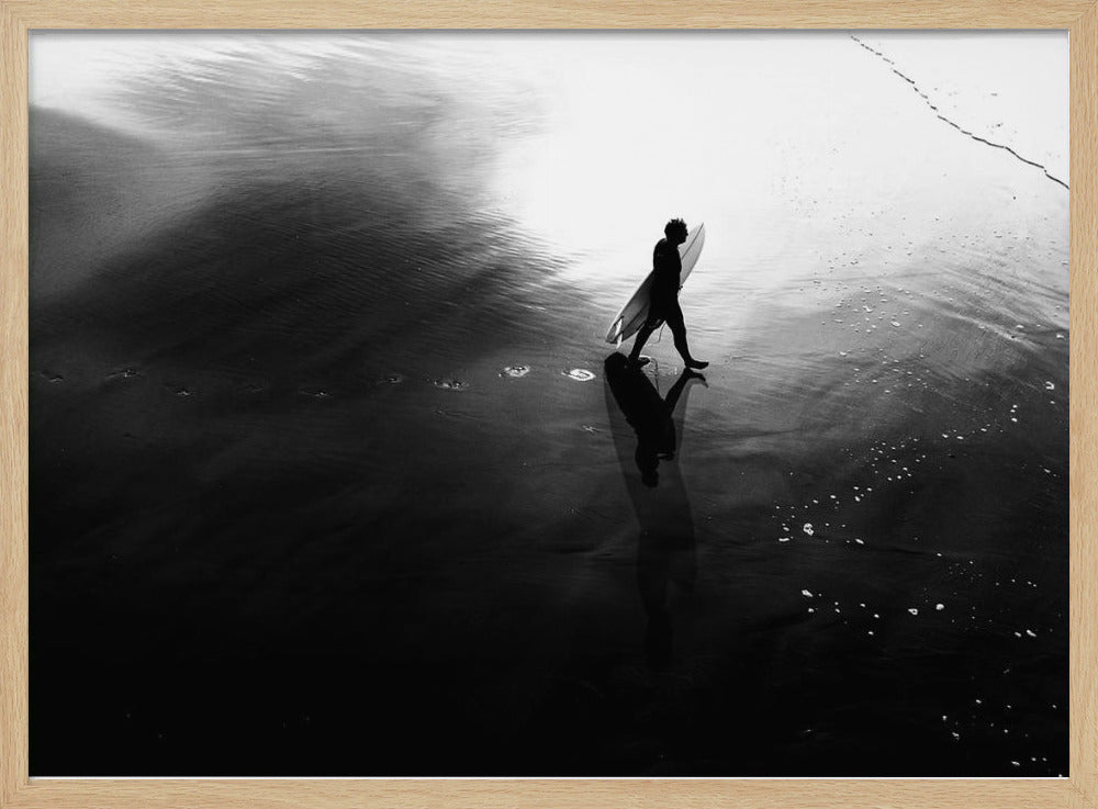 A high-contrast, black and white photograph of a surfer silhouetted against the bright reflection of the sun on the water. The surfer is carrying a surfboard and walking along the wet sand, leaving footprints behind. Their long shadow is cast in front of them on the dark, glistening sand. Print