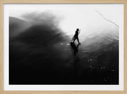 A high-contrast, black and white photograph of a surfer silhouetted against the bright reflection of the sun on the water. The surfer is carrying a surfboard and walking along the wet sand, leaving footprints behind. Their long shadow is cast in front of them on the dark, glistening sand. Print