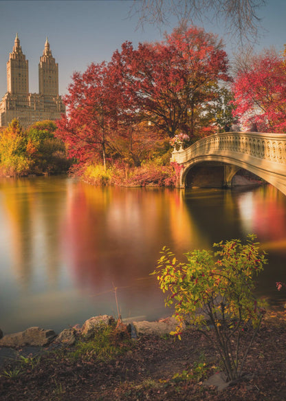 A picturesque autumn scene in Central Park featuring the Bow Bridge over a calm lake reflecting the vibrant red and yellow fall foliage, with the twin towers of The San Remo building visible in the background against a clear blue sky. Decor