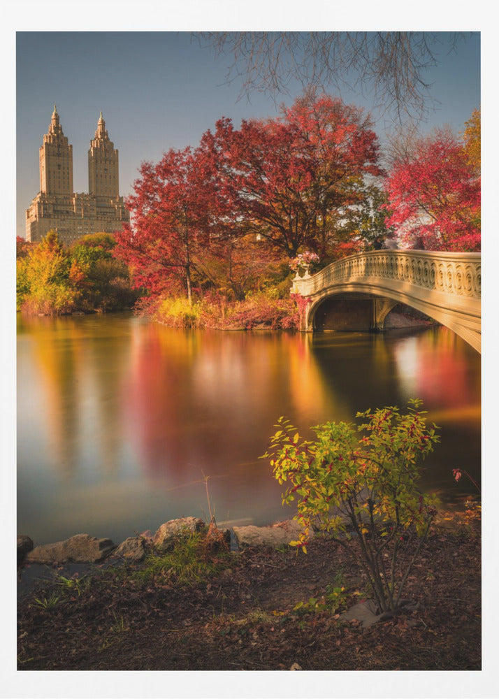 A picturesque autumn scene in Central Park featuring the Bow Bridge over a calm lake reflecting the vibrant red and yellow fall foliage, with the twin towers of The San Remo building visible in the background against a clear blue sky. Decor
