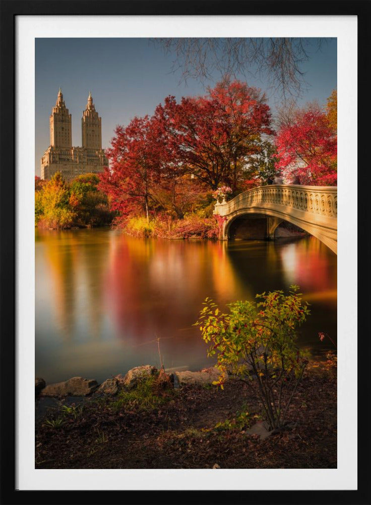 A picturesque autumn scene in Central Park featuring the Bow Bridge over a calm lake reflecting the vibrant red and yellow fall foliage, with the twin towers of The San Remo building visible in the background against a clear blue sky. Decor
