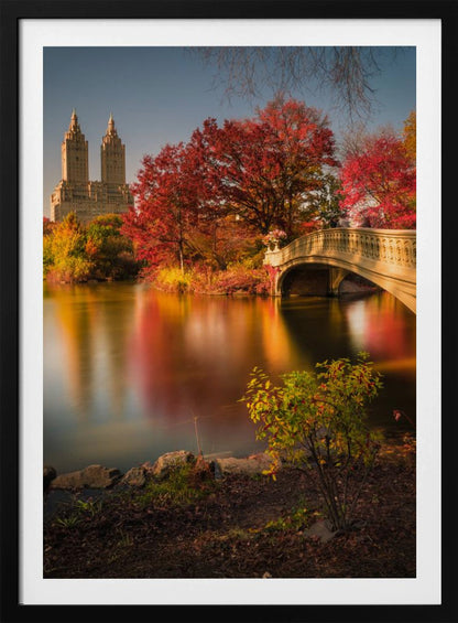 A picturesque autumn scene in Central Park featuring the Bow Bridge over a calm lake reflecting the vibrant red and yellow fall foliage, with the twin towers of The San Remo building visible in the background against a clear blue sky. Decor