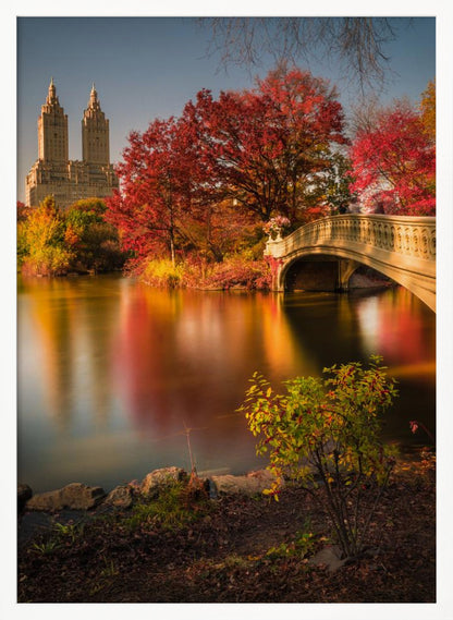 A picturesque autumn scene in Central Park featuring the Bow Bridge over a calm lake reflecting the vibrant red and yellow fall foliage, with the twin towers of The San Remo building visible in the background against a clear blue sky. Decor