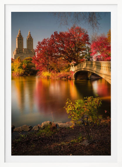 A picturesque autumn scene in Central Park featuring the Bow Bridge over a calm lake reflecting the vibrant red and yellow fall foliage, with the twin towers of The San Remo building visible in the background against a clear blue sky. Decor