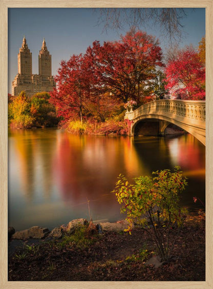 A picturesque autumn scene in Central Park featuring the Bow Bridge over a calm lake reflecting the vibrant red and yellow fall foliage, with the twin towers of The San Remo building visible in the background against a clear blue sky. Decor