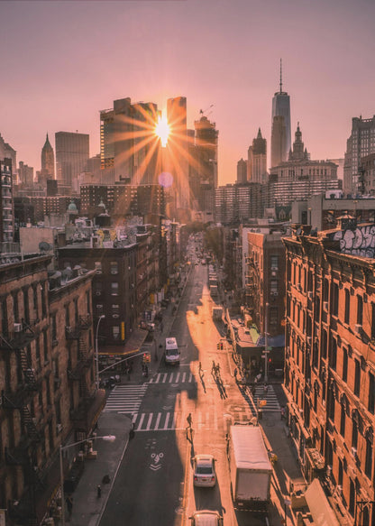 A high-angle view of a New York City street at sunset, with the sun bursting between skyscrapers and casting long golden shadows over the buildings and traffic below. Decor
