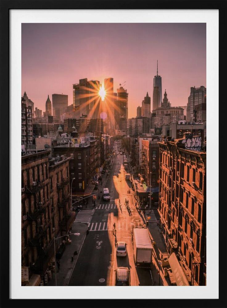 A high-angle view of a New York City street at sunset, with the sun bursting between skyscrapers and casting long golden shadows over the buildings and traffic below. Decor