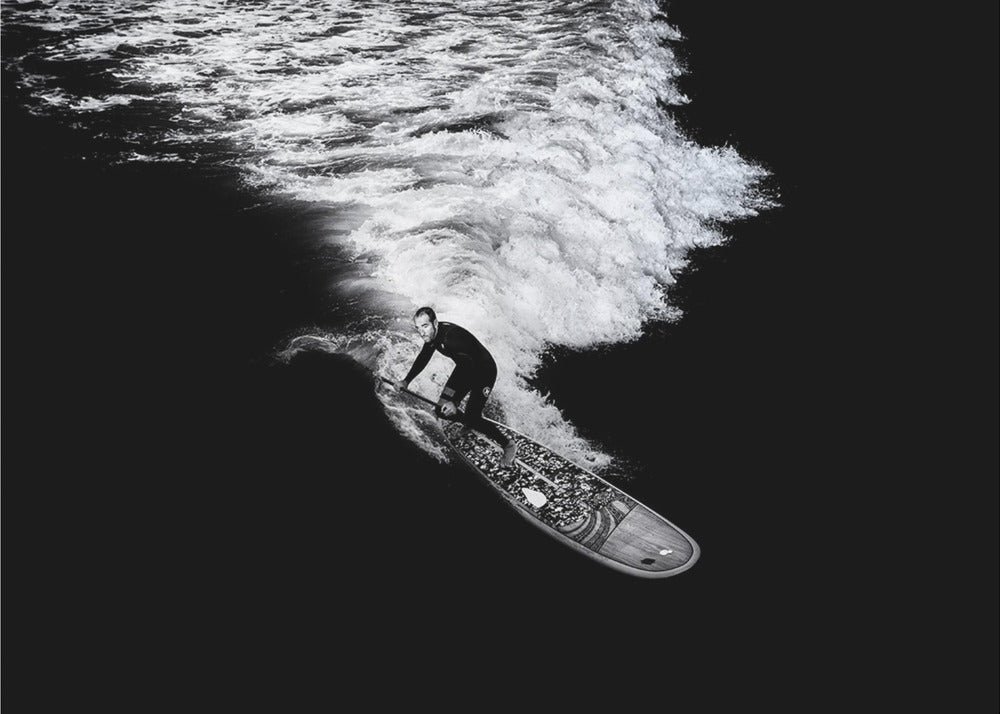 A dramatic black and white high-angle photograph of a man in a wetsuit paddleboarding on a wave, creating a stark white wake against the deep black water, all enclosed in a silver frame. Print