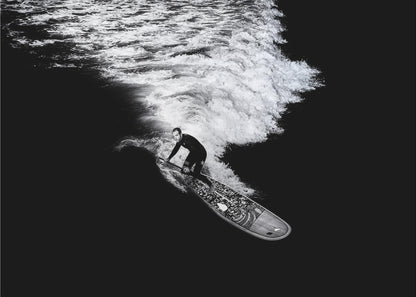 A dramatic black and white high-angle photograph of a man in a wetsuit paddleboarding on a wave, creating a stark white wake against the deep black water, all enclosed in a silver frame. Print