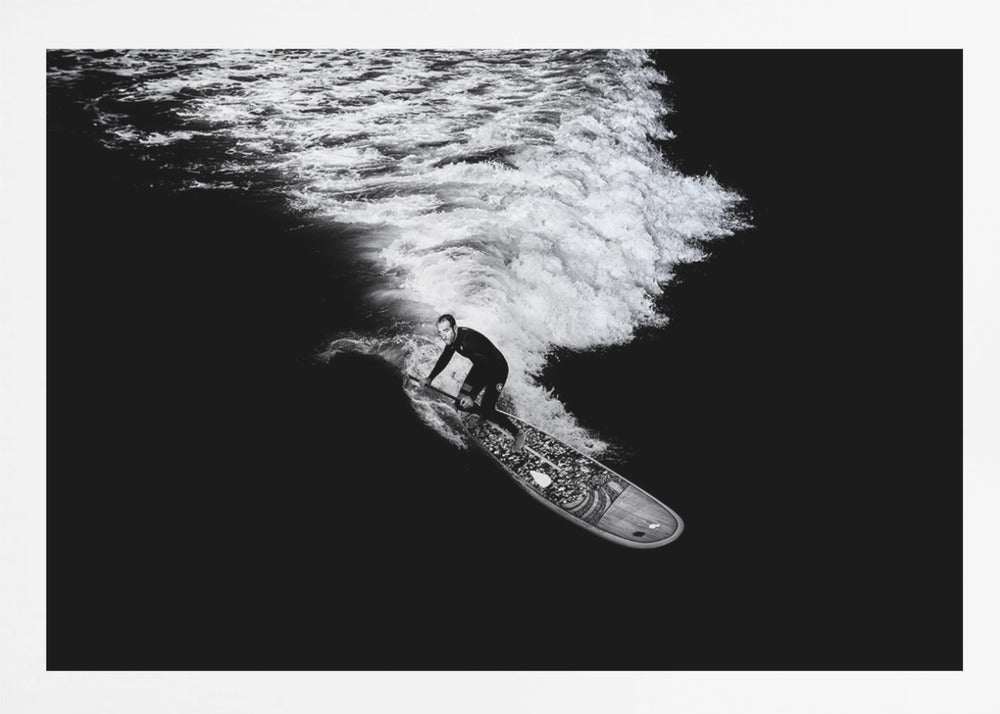 A dramatic black and white high-angle photograph of a man in a wetsuit paddleboarding on a wave, creating a stark white wake against the deep black water, all enclosed in a silver frame. Print