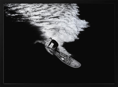 A dramatic black and white high-angle photograph of a man in a wetsuit paddleboarding on a wave, creating a stark white wake against the deep black water, all enclosed in a silver frame. Print