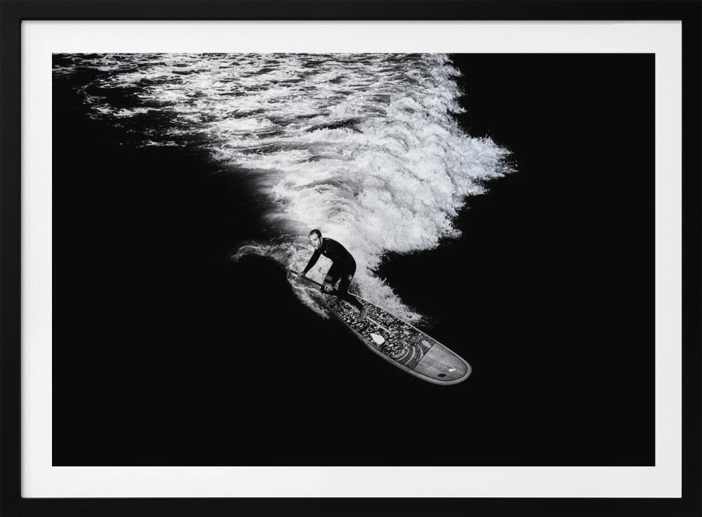 A dramatic black and white high-angle photograph of a man in a wetsuit paddleboarding on a wave, creating a stark white wake against the deep black water, all enclosed in a silver frame. Print