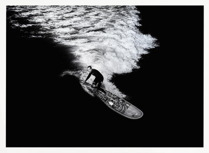 A dramatic black and white high-angle photograph of a man in a wetsuit paddleboarding on a wave, creating a stark white wake against the deep black water, all enclosed in a silver frame. Print