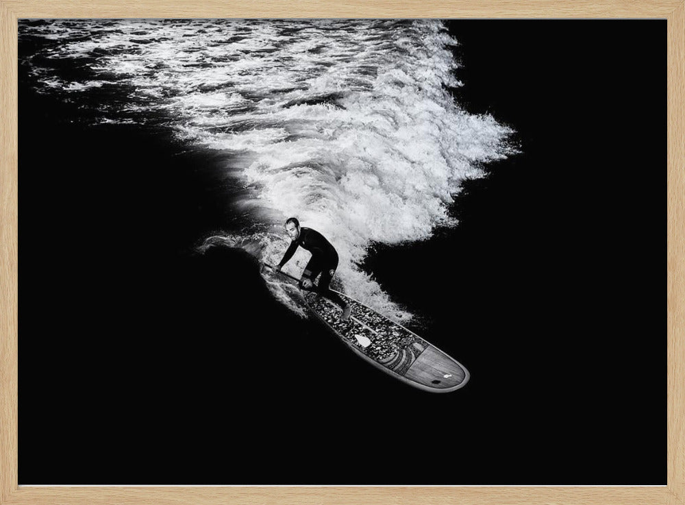 A dramatic black and white high-angle photograph of a man in a wetsuit paddleboarding on a wave, creating a stark white wake against the deep black water, all enclosed in a silver frame. Print