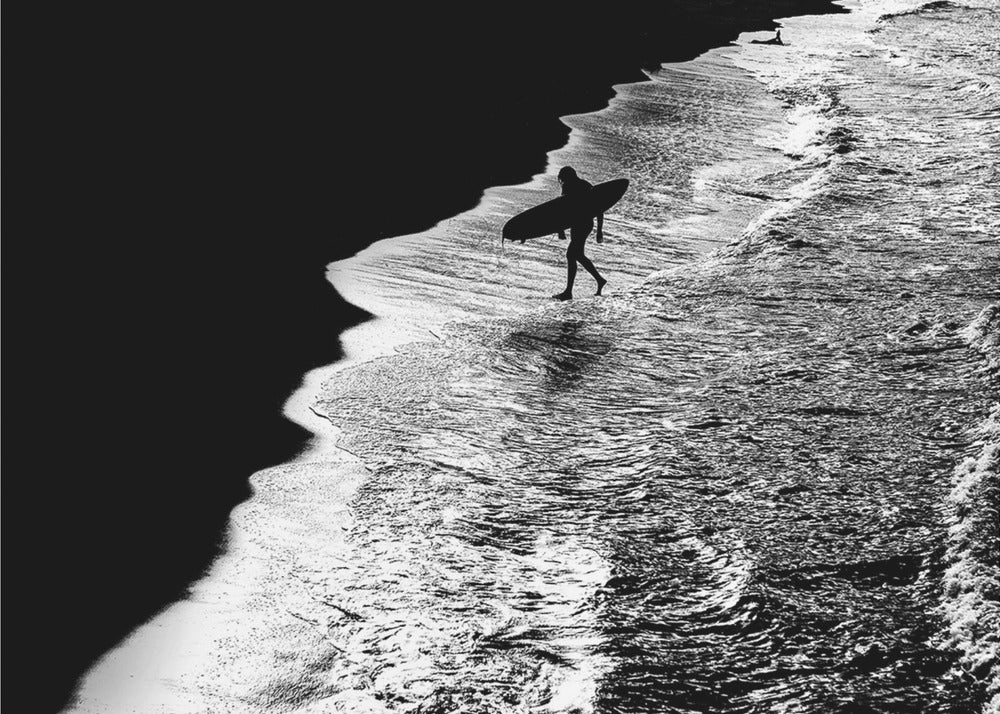 A high-contrast, black and white aerial photograph of a silhouetted surfer carrying a surfboard and walking along the shoreline. The image is divided diagonally, with the dark beach on the left and the white, foamy ocean waves on the right, creating a dramatic and graphic composition. The image is enclosed in a silver frame. Wall Art
