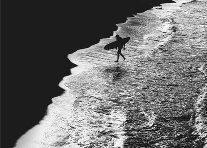 A high-contrast, black and white aerial photograph of a silhouetted surfer carrying a surfboard and walking along the shoreline. The image is divided diagonally, with the dark beach on the left and the white, foamy ocean waves on the right, creating a dramatic and graphic composition. The image is enclosed in a silver frame. Wall Art