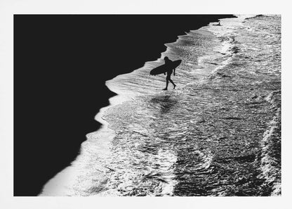 A high-contrast, black and white aerial photograph of a silhouetted surfer carrying a surfboard and walking along the shoreline. The image is divided diagonally, with the dark beach on the left and the white, foamy ocean waves on the right, creating a dramatic and graphic composition. The image is enclosed in a silver frame. Wall Art