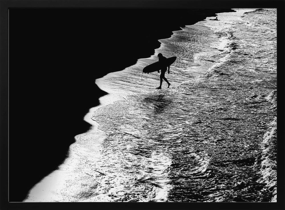 A high-contrast, black and white aerial photograph of a silhouetted surfer carrying a surfboard and walking along the shoreline. The image is divided diagonally, with the dark beach on the left and the white, foamy ocean waves on the right, creating a dramatic and graphic composition. The image is enclosed in a silver frame. Wall Art