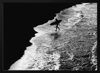 A high-contrast, black and white aerial photograph of a silhouetted surfer carrying a surfboard and walking along the shoreline. The image is divided diagonally, with the dark beach on the left and the white, foamy ocean waves on the right, creating a dramatic and graphic composition. The image is enclosed in a silver frame. Wall Art