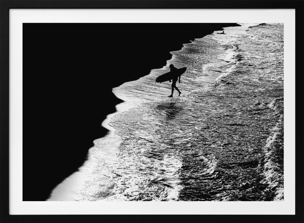 A high-contrast, black and white aerial photograph of a silhouetted surfer carrying a surfboard and walking along the shoreline. The image is divided diagonally, with the dark beach on the left and the white, foamy ocean waves on the right, creating a dramatic and graphic composition. The image is enclosed in a silver frame. Wall Art
