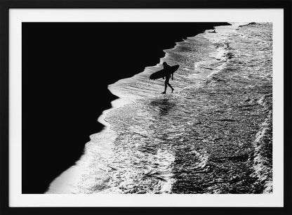 A high-contrast, black and white aerial photograph of a silhouetted surfer carrying a surfboard and walking along the shoreline. The image is divided diagonally, with the dark beach on the left and the white, foamy ocean waves on the right, creating a dramatic and graphic composition. The image is enclosed in a silver frame. Wall Art