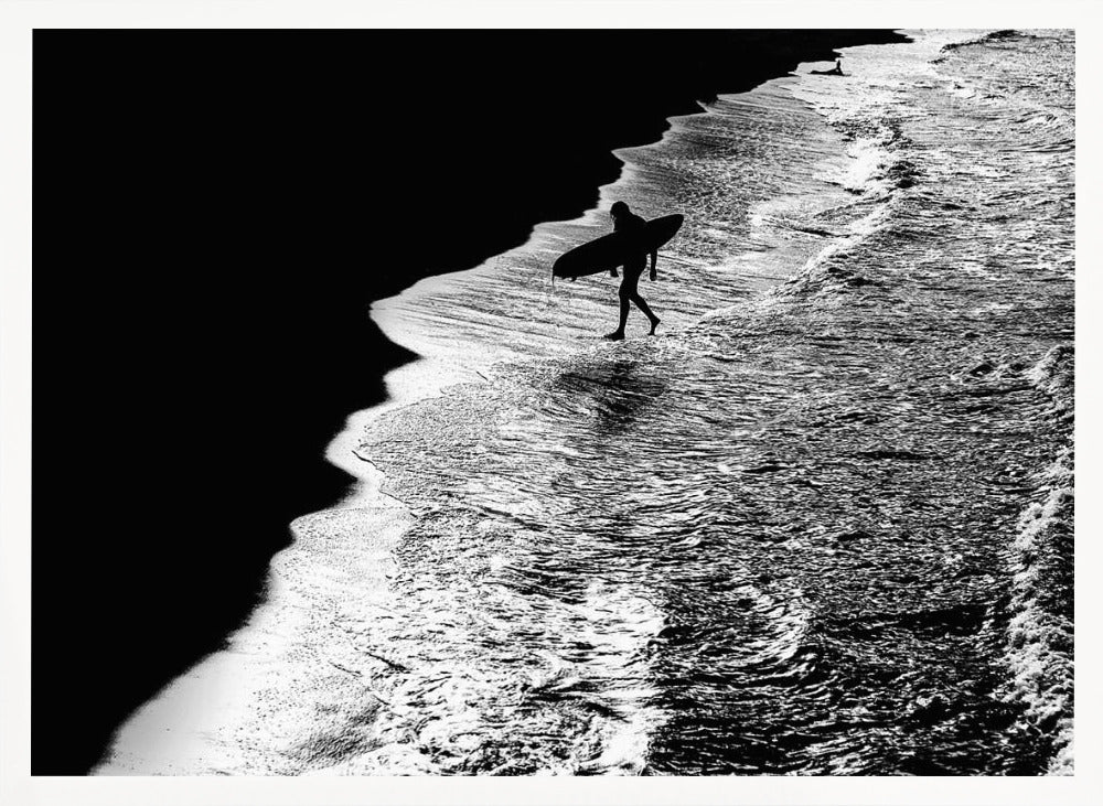 A high-contrast, black and white aerial photograph of a silhouetted surfer carrying a surfboard and walking along the shoreline. The image is divided diagonally, with the dark beach on the left and the white, foamy ocean waves on the right, creating a dramatic and graphic composition. The image is enclosed in a silver frame. Wall Art