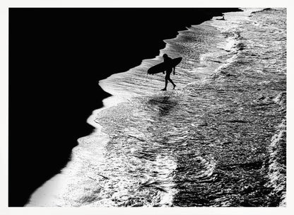 A high-contrast, black and white aerial photograph of a silhouetted surfer carrying a surfboard and walking along the shoreline. The image is divided diagonally, with the dark beach on the left and the white, foamy ocean waves on the right, creating a dramatic and graphic composition. The image is enclosed in a silver frame. Wall Art