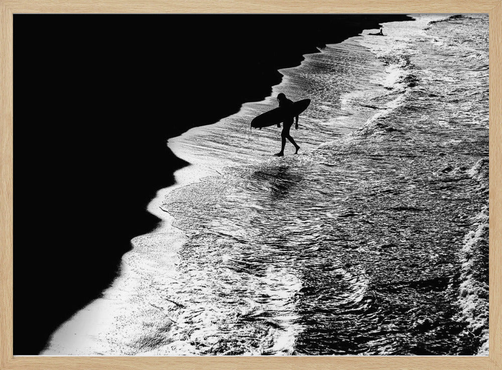 A high-contrast, black and white aerial photograph of a silhouetted surfer carrying a surfboard and walking along the shoreline. The image is divided diagonally, with the dark beach on the left and the white, foamy ocean waves on the right, creating a dramatic and graphic composition. The image is enclosed in a silver frame. Wall Art