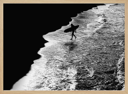 A high-contrast, black and white aerial photograph of a silhouetted surfer carrying a surfboard and walking along the shoreline. The image is divided diagonally, with the dark beach on the left and the white, foamy ocean waves on the right, creating a dramatic and graphic composition. The image is enclosed in a silver frame. Wall Art