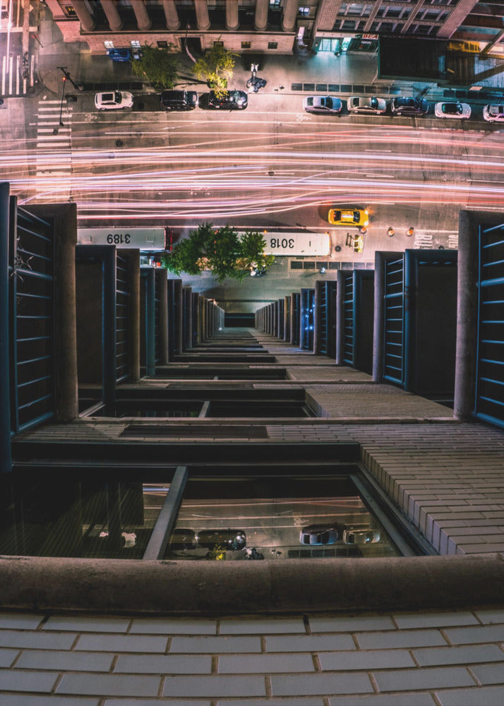 A dizzying bird's-eye view looking straight down the side of a skyscraper into a busy city street at night, with long exposure light trails from traffic creating streaks of red and white below. Wall Art