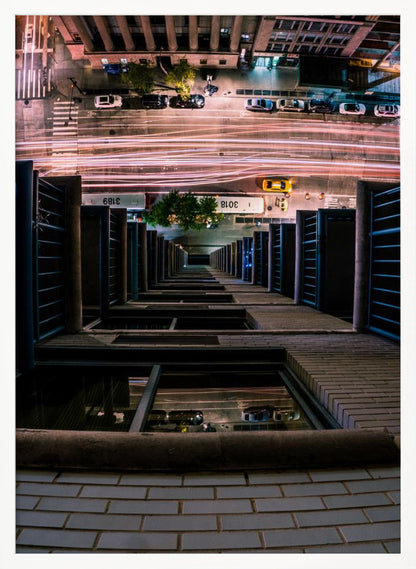 A dizzying bird's-eye view looking straight down the side of a skyscraper into a busy city street at night, with long exposure light trails from traffic creating streaks of red and white below. Wall Art