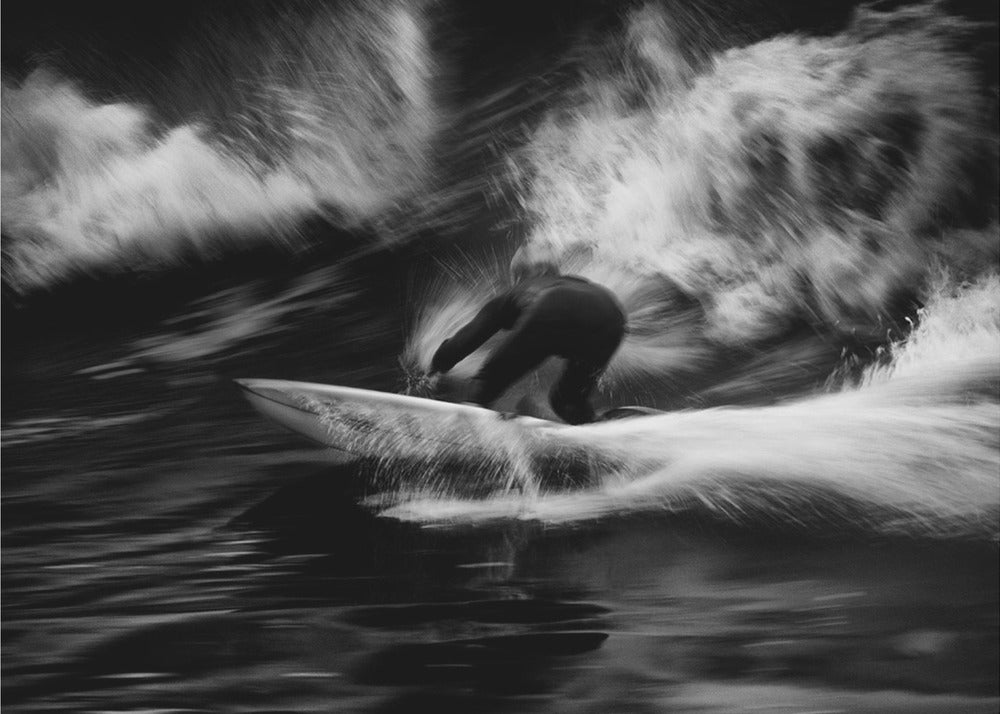 A dynamic black and white action photograph of a surfer in a dark wetsuit, crouched low on a surfboard while riding a powerful wave. Motion blur captures the speed and energy of the moment, with white water spray exploding around the surfer against the dark ocean. Print