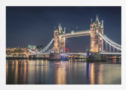 A stunning night photograph of London's iconic Tower Bridge, fully illuminated with golden and white lights that cast a shimmering reflection on the dark waters of the River Thames below, all set against a deep night sky and enclosed in a silver frame. Poster