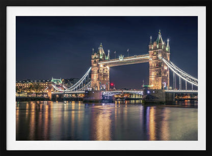 A stunning night photograph of London's iconic Tower Bridge, fully illuminated with golden and white lights that cast a shimmering reflection on the dark waters of the River Thames below, all set against a deep night sky and enclosed in a silver frame. Poster