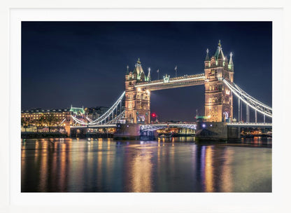 A stunning night photograph of London's iconic Tower Bridge, fully illuminated with golden and white lights that cast a shimmering reflection on the dark waters of the River Thames below, all set against a deep night sky and enclosed in a silver frame. Poster