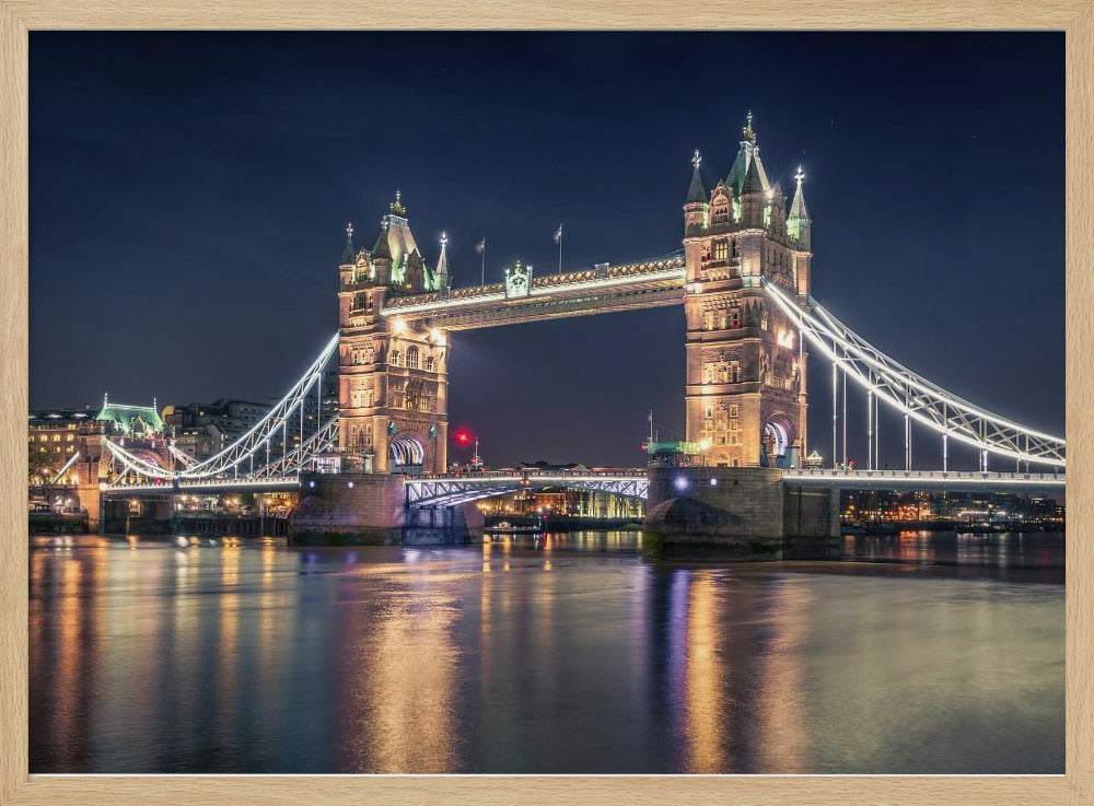 A stunning night photograph of London's iconic Tower Bridge, fully illuminated with golden and white lights that cast a shimmering reflection on the dark waters of the River Thames below, all set against a deep night sky and enclosed in a silver frame. Poster